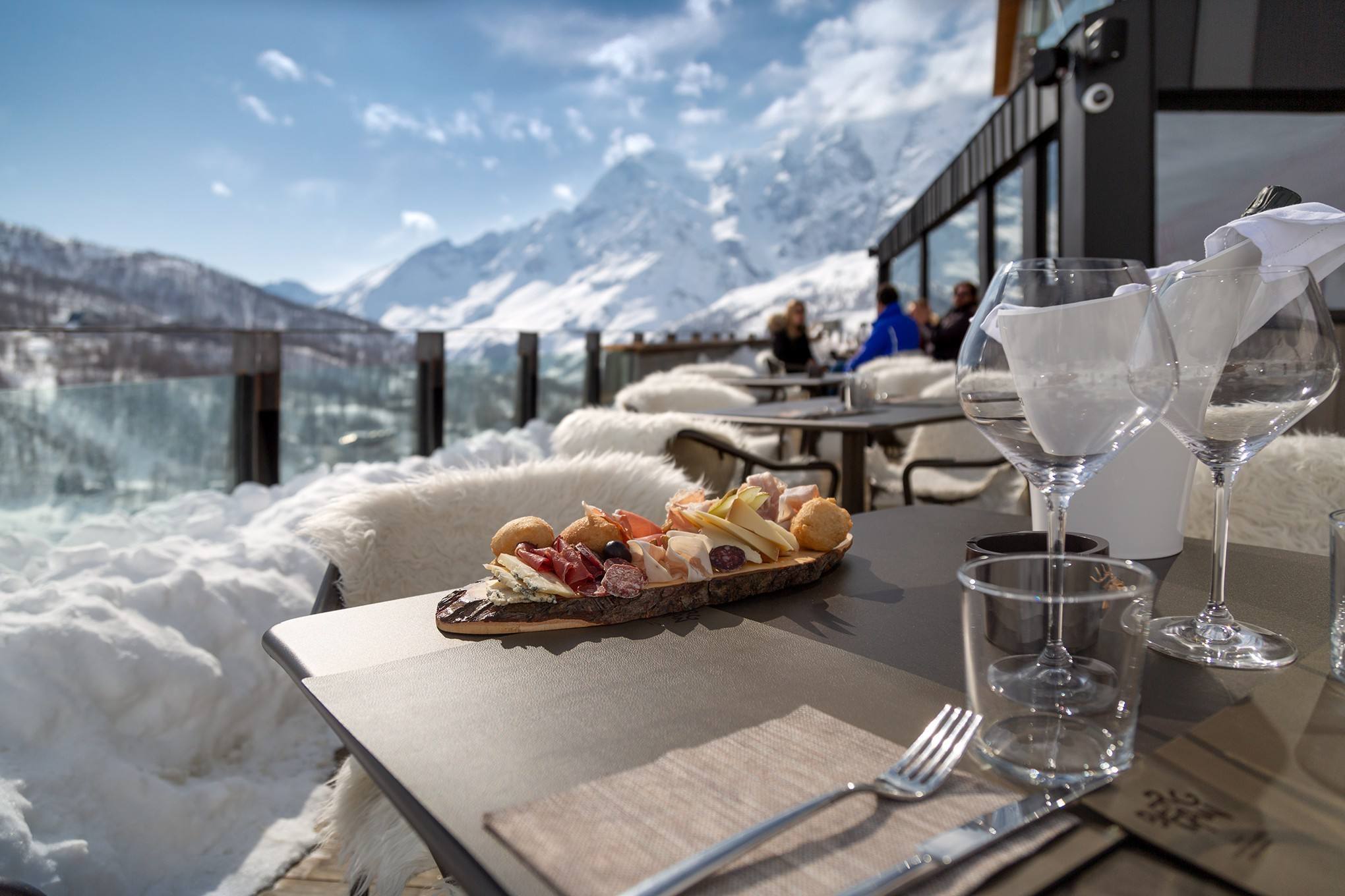 Tagliere e calici sulla terrazza innevata, con ospiti sullo sfondo